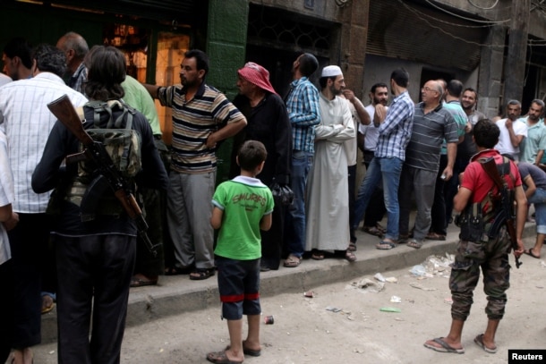 Rebel fighters stand guard as people queue for bread in the rebel held al-Shaar neighborhood of Aleppo, Syria July 14, 2016.