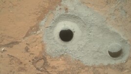 Na jednoj od fotografija koju je Kjuriositi poslao NASI na zemlju vidi se stena poznata kao Džon Klajn sa koje je rover uzeo prve uzorke tla Marsa.   