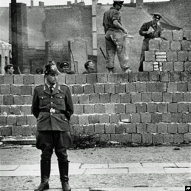 Một cảnh vệ Tây Berlin đứng trước bức tường bê tông chia cắt Đông và Tây Berlin ở Bernauer Strasse, trong khi các công nhân Đông Berlin chất các khối gạch lên bức tường để xây cao bức tường chắn này, 7/10/1961.