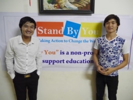 Phạm Minh Dap (trái) và em trai đứng trước một tấm băng rôn quảng cáo lớp học ngoại ngữ miễn phí Stand By You cho học sinh, sinh viên nghèo (Marianne Brown/VOA)