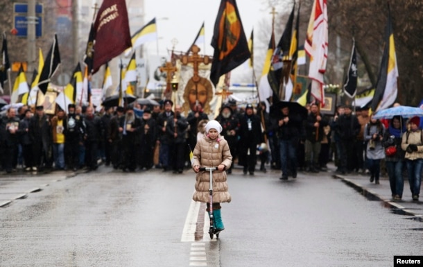 FILE - A girl rides a kick scooter as people walk during a "Russian March" demonstration, organized by the "Russian Coalition of Action" movement, seen in the background, on National Unity Day in Moscow, Nov. 4, 2013.