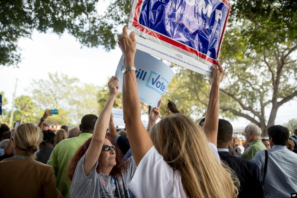 两派标语对峙。希拉里&middot;克林顿在一个提前投票地点外欢迎支持者的时候，一名克林顿的支持者试图挡住一名川普支持者举起的标语牌