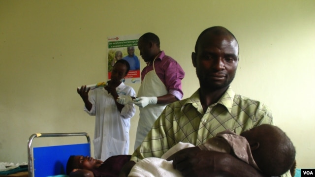 Savio Niwagaba holds his newborn baby as his wife Chrisente, behind him, has a contraceptive implant inserted in her arm, Kanungu, Uganda, June 19, 2012. (VOA / Hilary Heuler)