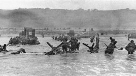 Some of the first assault troops to hit the beach of Normandy, France, on June 6, 1944 take cover behind enemy obstacles to fire on German forces as others follow the first tanks through the water toward the German-held shore during World War II
