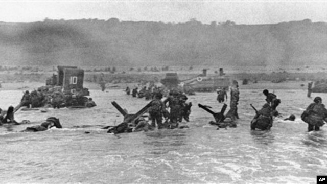 Some of the first assault troops to hit the beach of Normandy, France, on June 6, 1944 take cover behind enemy obstacles to fire on German forces as others follow the first tanks through the water toward the German-held shore during World War II