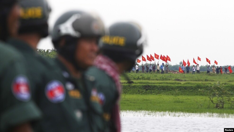 FILE - Cambodian police officers stand guard as Vietnamese people wave their national flags at the Cambodia-Vietnam border in Svay Rieng province, July 19, 2015. 