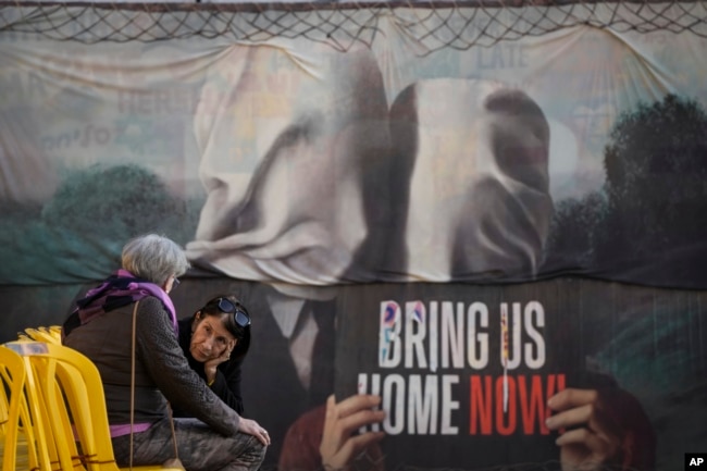 Dua perempuan berbincang di samping mural yang menuntut pemulangan sandera yang ditawan Hamas di Jalur Gaza, di Tel Aviv, Israel, Sabtu, 18 Januari 2025. (Foto: AP/Oded Balilty)