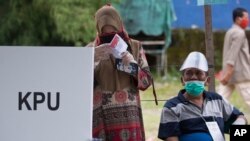 Seorang perempuan mengenakan masker dan sarung tangan plastik tengah memeriksa surat suaranya disaksikan petugas pemungutan suara dalam PIlkada di sebuah TPS di Medan, Sumatera Utara, 9 Desember 2020. (AP Photo/Binsar Bakkara)