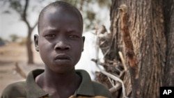 In this photo taken Saturday, Feb. 15, 2014, Ajing Abiik, 12, who fled from his hometown of Bor when the fighting broke out in December, stands next to the tree where he and his two brothers sleep in Minkaman IDP camp, Awerial County, in South Sudan.