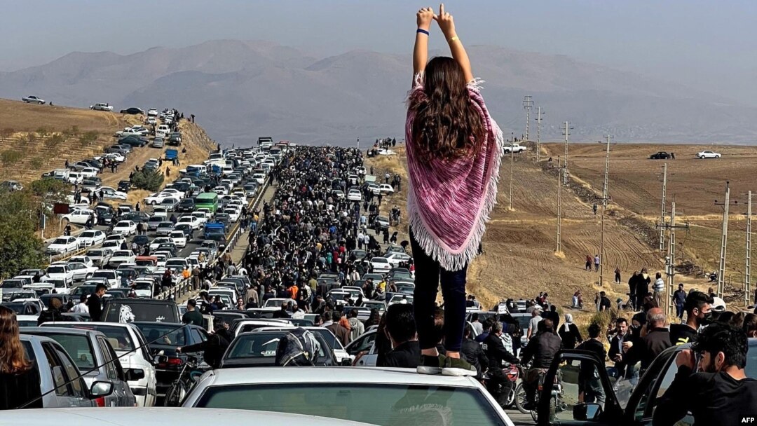 Seorang perempuan tanpa mengenakan hijab berdiri di atas mobil sementara ribuan pedemo berjalan ke arah makam di kota kelahiran Mahsa Amini untuk memperingati 40 hari wafatnya perempuan Iran itu, 26 Oktober 2022. (Foto: UGC via AP)