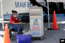 In this Aug. 7, 2020, photo, a poll worker monitors a ballot drop box, identified in English, Spanish and Haitian Creole, for mail-in ballots outside of a polling station during early voting in Miami Beach, Florida.