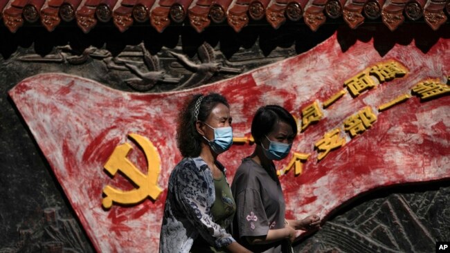 Women wearing face masks walk by a mural depicting a communist party flag and a party slogan in Beijing, Tuesday, Sept. 6, 2022. (AP Photo/Andy Wong)