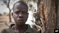 In this photo taken Saturday, Feb. 15, 2014, Ajing Abiik, 12, who fled from his hometown of Bor when the fighting broke out in December, stands next to the tree where he and his two brothers sleep in Minkaman IDP camp, Awerial County, in South Sudan. The fighting in the world’s newest country has left thousands of its youngest citizens either orphans or separated from their parents, increasing their vulnerability to sickness, malnutrition and recruitment by warring groups as child soldiers. (AP Photo/Mackenzie Knowles-Coursin)