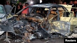 People stand near a damaged vehicle at the site of a bomb blast, outside the Sayeda Zeinab shrine city south of the Syrian capital Damascus, Syria, July 27, 2023. (SANA/Handout via Reuters)