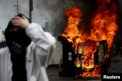 A masked protester stands in front of a burning car during clashes at a demonstration as part of the 10th day of nationwide strikes and protests against French government's pension reform in Nantes, France, March 28, 2023.