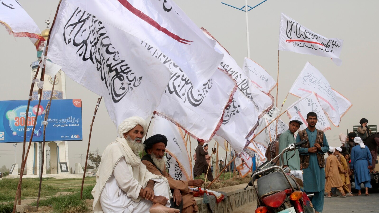 Sejumlah warga berada di pinggir jalan yang dihiasi bendera Taliban di Kabul, Afghanistan, pada 15 Agustus 2023. Bendera tersebut dipasang sebagai perayaan dua tahun kembalinya Taliban berkuasa di negara itu. (Foto: AP/Abdul Khaliq)