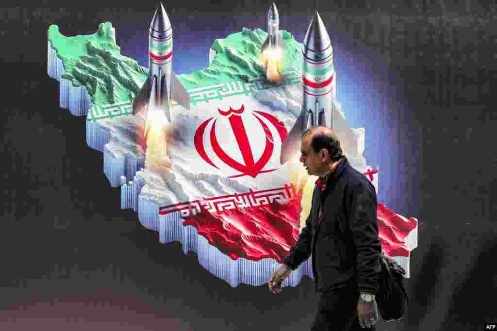 Seorang pria berjalan melewati spanduk yang menggambarkan peluncuran rudal (dalam serangan ke Israel) dan peta negara Iran yang diwarnai dengan bendera Iran, di pusat ibu kota Teheran. (AFP)&nbsp;