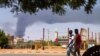 Men walk down a street in Khartoum on June 8, 2023, as smoke rises from behind buildings amid continuing fighting between the army and paramilitary forces. 