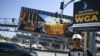 Seorang pendukung serikat pekerja seni, SAG AFTRA, mengacungkan poster bertuliskan "SAF AFTRA Dukung WGA" dalam demo para penulis di luar Universal Studios Hollywood di Los Angeles, California, Jumat, 30 Juni 2023. (Foto: Robyn Beck/AFP)