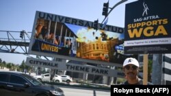 Seorang pendukung serikat pekerja seni, SAG AFTRA, mengacungkan poster bertuliskan "SAF AFTRA Dukung WGA" dalam demo para penulis di luar Universal Studios Hollywood di Los Angeles, California, Jumat, 30 Juni 2023. (Foto: Robyn Beck/AFP)