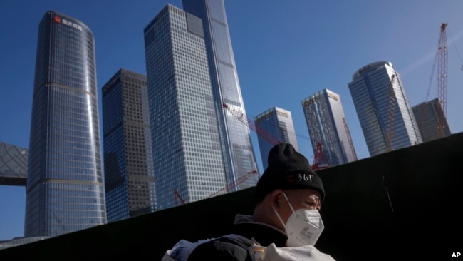 FILE - A migrant worker wearing a face mask walks by construction cranes near the office buildings at the central business district in Beijing, March 15, 2023.