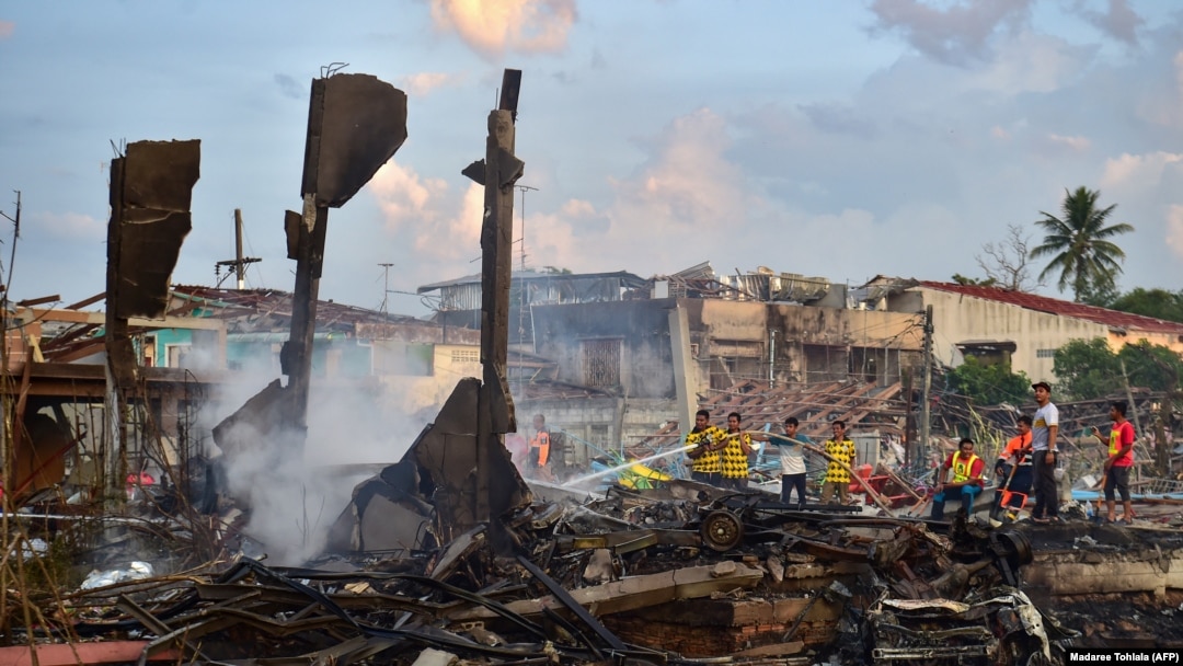 Petugas pemadam kebakaran memadamkan bara api di rumah-rumah yang hancur setelah ledakan di sebuah gudang penyimpanan kembang api hingga menewaskan 9 orang dan melukai lebih dari 100 orang lainnya di distrik Sungai Kolok, Thailand, Sabtu, 29 Juli 2023. (Foto: Madaree Tohlala/AFP)