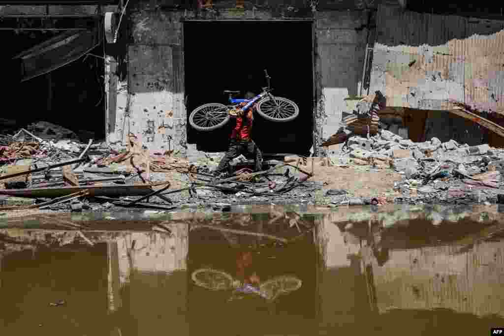 A Palestinian youth carries his bike as he walks on the rubble of destroyed buildings along the edge of a pool of stagnant water in Khan Yunis in the southern Gaza Strip, as the conflict between Israel and the Palestinian militant group Hamas continues.