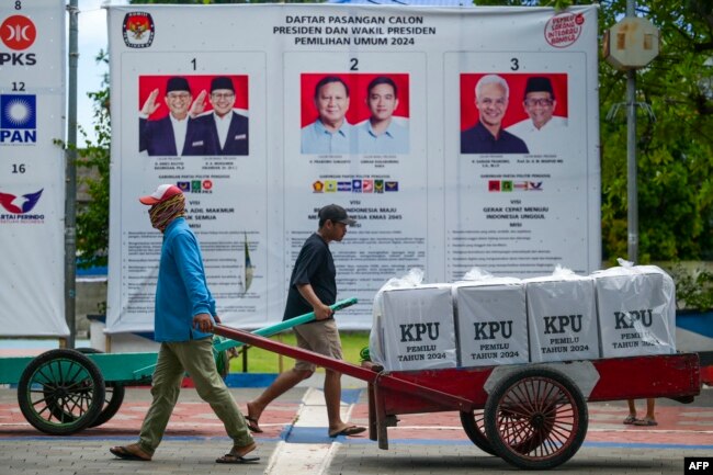 Petugas membawa kotak suara melewati poster calon pemilu setelah diangkut dengan perahu motor ke Kepulauan Seribu di Jakarta, 9 Februari 2024, menjelang pemilihan presiden dan legislatif Indonesia yang dijadwalkan digelar pada 14 Februari. (BAY ISMOYO / AFP)