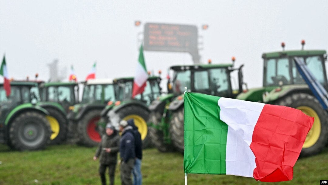 Bendera nasional Italia berkibar di depan traktor yang diparkir di sepanjang jalan saat para petani melakukan aksi protes di dekat pintu masuk jalan raya di Melegnano, dekat Milan, 30 Januari 2023. (GABRIEL BOUYS / AFP)