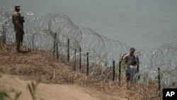 Migrants walk along concertina wire past a guardsman after crossing the Rio Grande from Mexico near the site where large buoys are being deployed to be used as a border barrier in Eagle Pass, Texas, July 12, 2023.