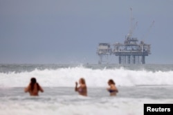 Orang-orang berenang di depan anjungan anjungan minyak lepas pantai di Huntington Beach, California, AS 4 Juli 2024. (REUTERS/Etienne Laurent)