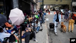 FILE - Migrants sit in a queue outside the Roosevelt Hotel, which is being used by the city as temporary housing, on July 31, 2023, in New York. New York mayor calls for changes in city’s migrant sanctuary status.