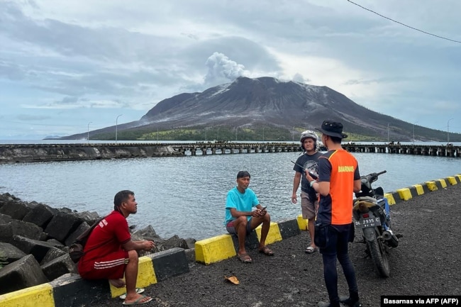 Seorang anggota Basarnas berbincang dengan warga di Sitaro, Sulawesi Utara, dengan latar belakang gunung berapi Gunung Ruang yang mengeluarkan asap. (Foto: Basarnas via AFP)