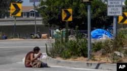 A homeless woman sits on the street at the entrance to an off-ramp to the CA-105 freeway next to a homeless encampment, July 26, 2024, in Los Angeles, California. 