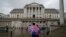 Seorang pengunjung dengan payung bermotif bendera Inggris berdiri di depan gedung bank sentral, Bank of England, di London, 3 November 2022. Bank of England akan menerapkan prosedur kepailitan kepada anak perusahaan Silicon Valley Bank di Inggris. (Foto: Kin Cheung/AP Photo)