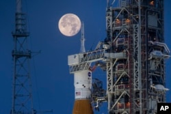 FILE - Bulan purnama terlihat di belakang Sistem Peluncuran Antariksa Artemis I (SLS) dan wahana antariksa Orion, di atas peluncur bergerak, di Pusat Antariksa Kennedy NASA di Florida pada 14 Juni 2022. (Cory Huston/NASA via AP)