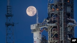 Bulan purnama terlihat di belakang Artemis I Space Launch System (SLS) dan pesawat ruang angkasa Orion di Kennedy Space Center NASA di Florida pada 14 Juni 2022. (Cory Huston/NASA via AP)