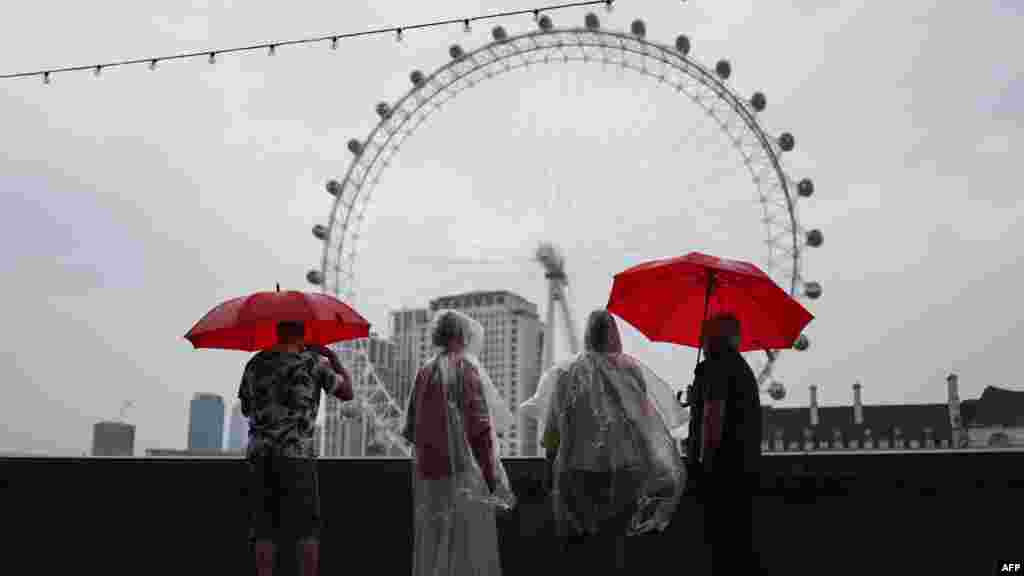 People shelter from the rain under umbrellas while looking at the London Eye, from Embankment by the River Thames, in central London, on a gloomy summer&#39;s day.