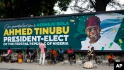 FILE - People sit under a billboard with a congratulatory message of the President-elect Bola Ahmed Tinubu of the All Progressives Congress in Lagos, Nigeria, March 5, 2023. 