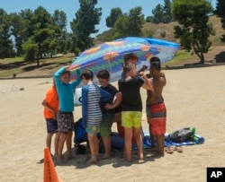 Teens seek shelter in the shade after being asked by lifeguards to retrieve their shoes before entering the sunny beach area to avoid burned feet at Castaic Lake beach as temperatures rise, July 8, 2024, in Castaic, California.
