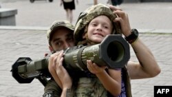 A Ukrainian serviceman helps a boy wearing body armor to hold an AT4 Swedish man-portable anti-tank weapon at a volunteers point in the center of Kyiv on Aug. 10, 2023.