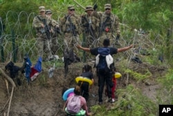 Seorang migran memberi isyarat kepada Pengawal Nasional Texas yang berdiri di belakang kawat berduri di tepi sungai Rio Grande, dilihat dari Matamoros, Meksiko, Kamis, 11 Mei 2023. (AP/Fernando Llano)