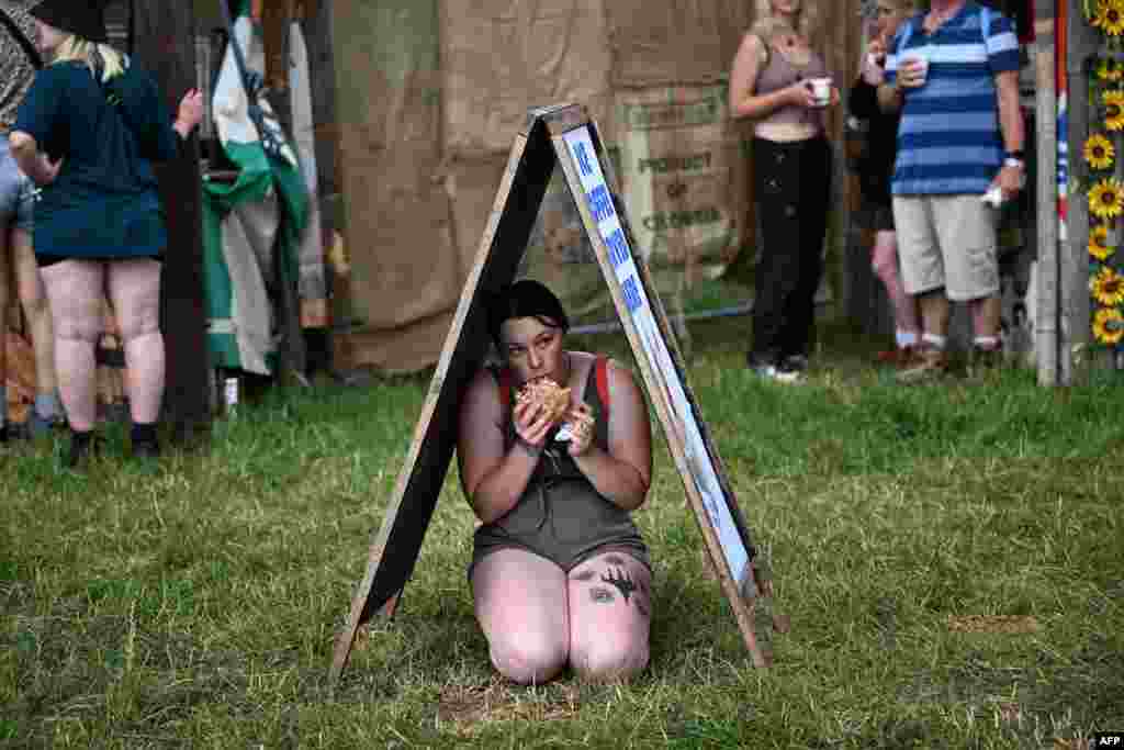 A festivalgoer eats while sheltering from the rain beneath an advertising board outside a food stall on the first day of the Glastonbury festival in the village of Pilton, in Somerset, southwest England.