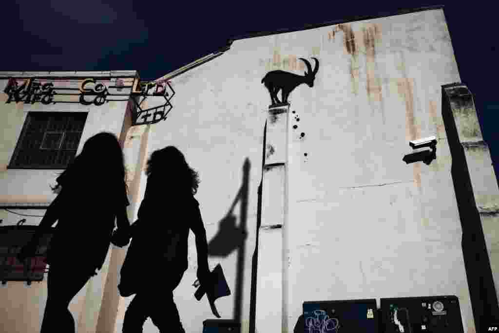 People walk past a stenciled Banksy artwork, depicting a mountain goat and falling rubble at the top of a pillar, on the side of a building in Kew Gardens, west London, Aug. 5, 2024.