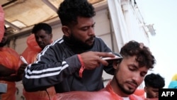 Migran asal Bangladesh tampak mencukur rambut rekannya saat berada di atas kapal penyelamat Ocean Viking menuyusul operasi penyelamatan yang dilakukan di lepas pantai Malta, pada 21 Mei 2024. (Foto: AFP/Clément Melki)