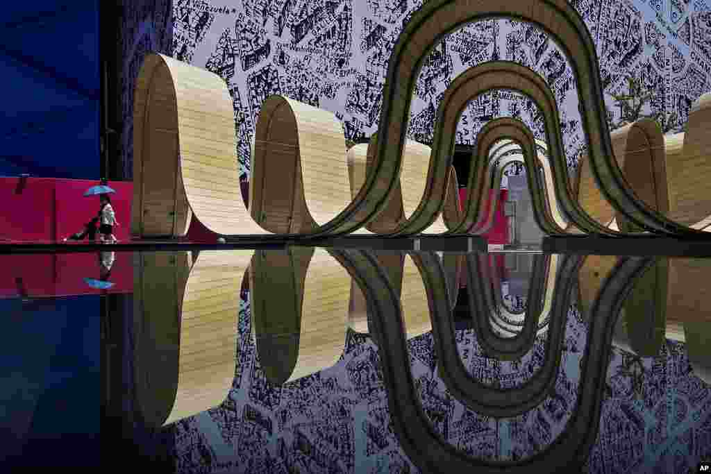 A couple carrying an umbrella walk with their child passing by a reflection of an art installation at an outdoor shopping mall in Beijing.