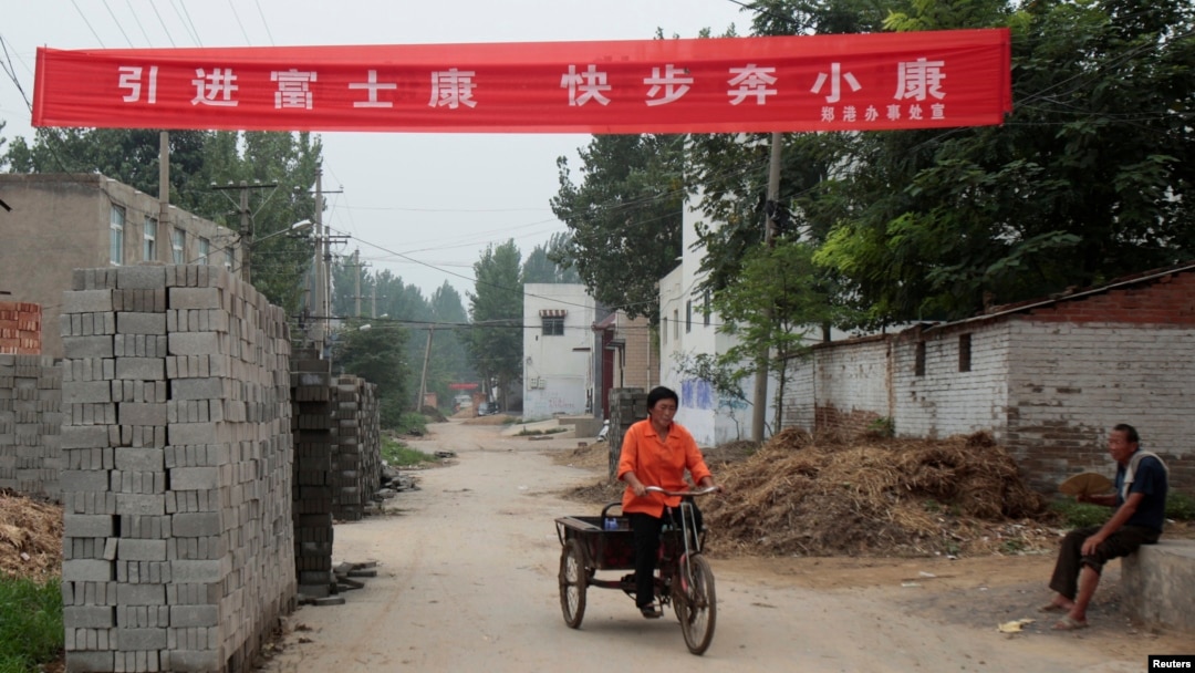 资料照：中国河南省郑州富士康工厂建筑工地旁的一个村庄，一名女子蹬着板车路过悬挂的“引进富士康，快步奔小康”的横幅标语。（2010年8月11日）