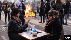 Two women have a drink on a cafe terrace as a garbage can is set ablaze nearby during a demonstration on the 11th day of protests against a pension reform program, in Paris, April 6, 2023.