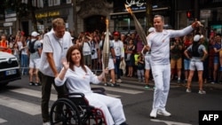 AFP Lebanese photographer Christina Assi (C) helped by AFP video journalist Dylan Collins (L), waves after receiving the Olympic flame during the Olympic Torch Relay in Vincennes, near Paris, July 21, 2024, ahead of the Paris 2024 Olympic Games.