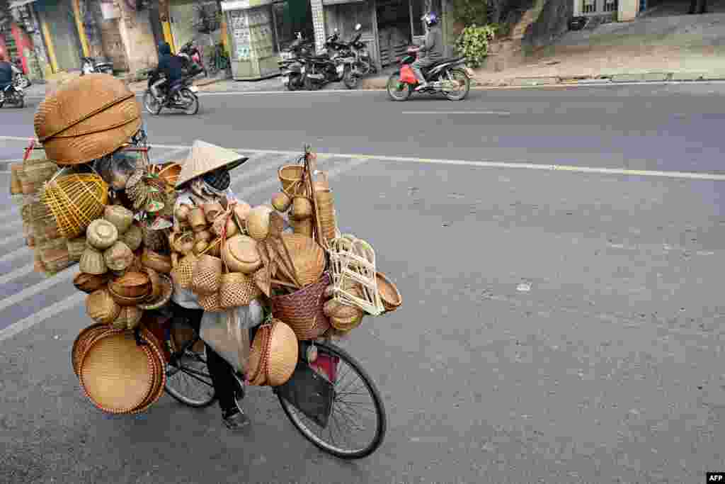 A woman rides a bicycle transporting bamboo and rattan household items for sale on a street in Hanoi, Vietnam.